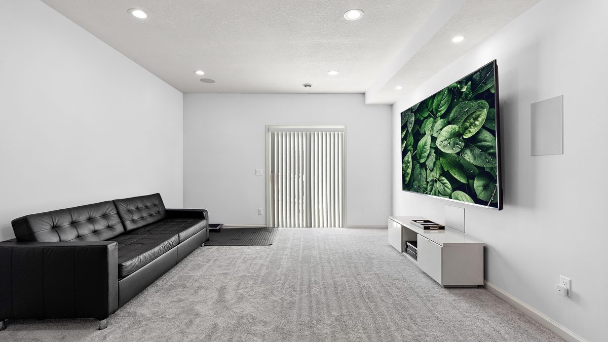 A finished basement media room with a wall-mounted screen, carpet, recessed lighting and a soffit running along one wall.