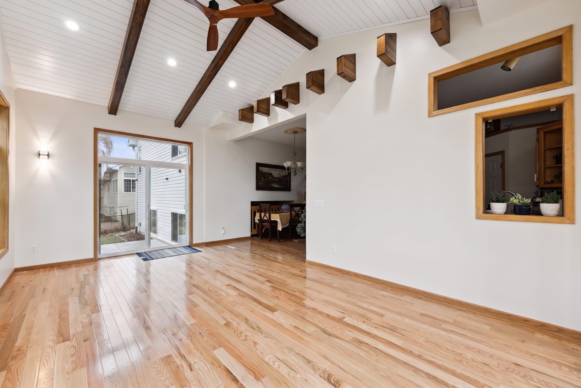 Vaulted great-room addition with dark stained beams, a ceiling fan and red oak flooring, open through to the dining area