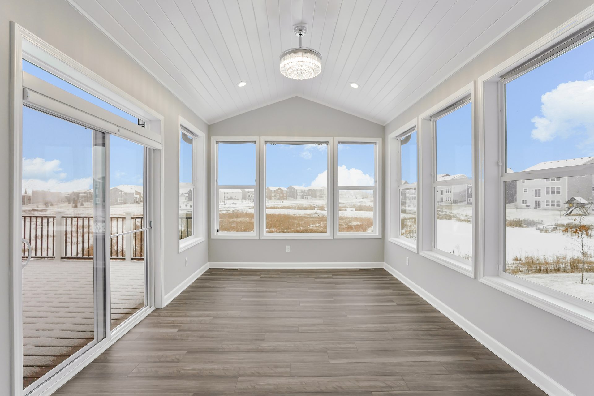 Four-season room with a vaulted shiplap ceiling, windows on three sides and gray plank flooring, looking out over snow