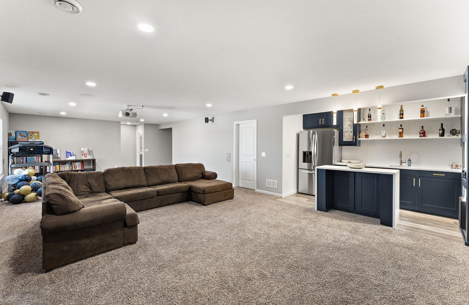 Finished basement family area with a brown sectional on carpet and a navy wet bar with open shelving beyond