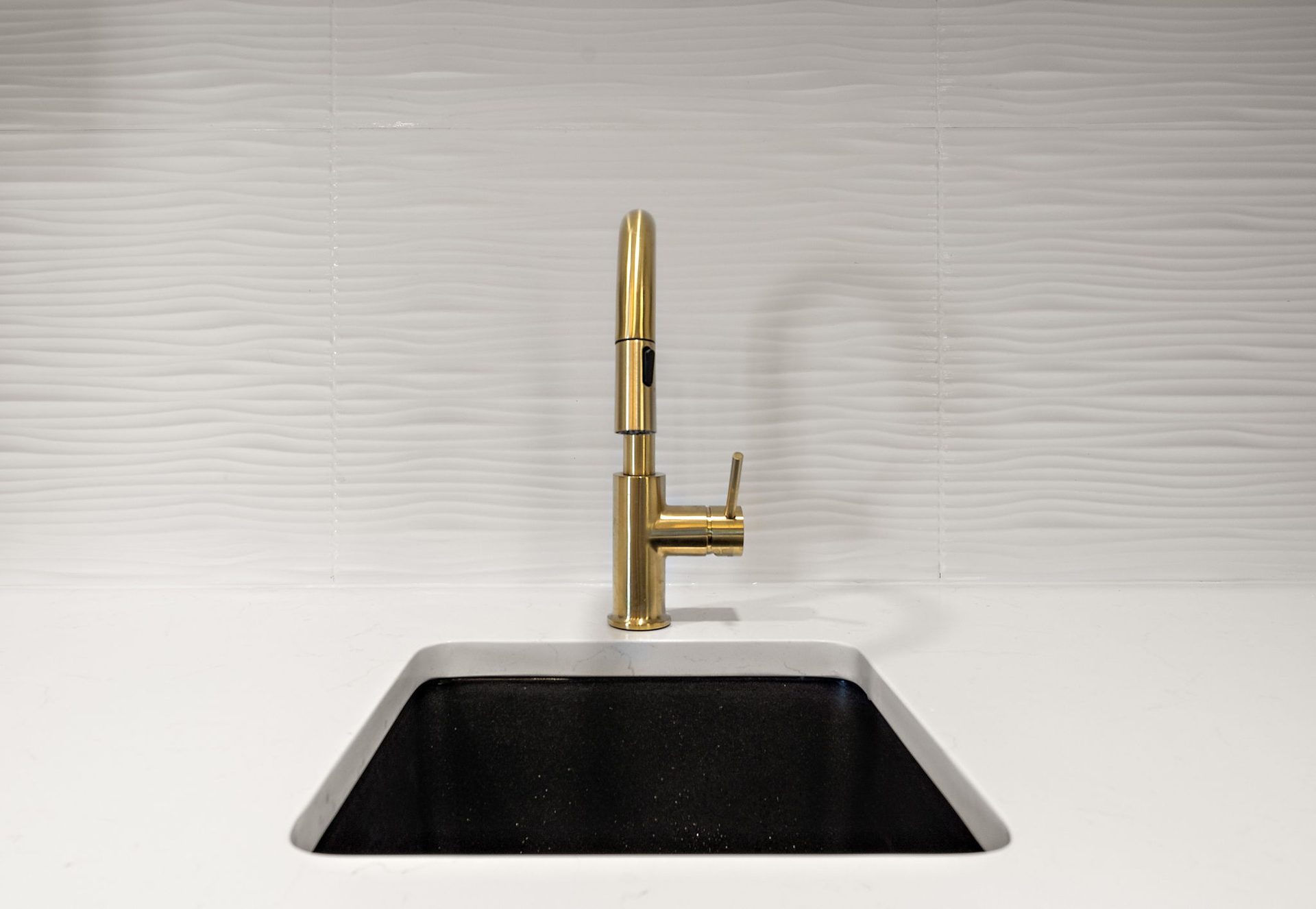 Bar sink detail with a brass faucet, undermount basin and a textured white tile backsplash