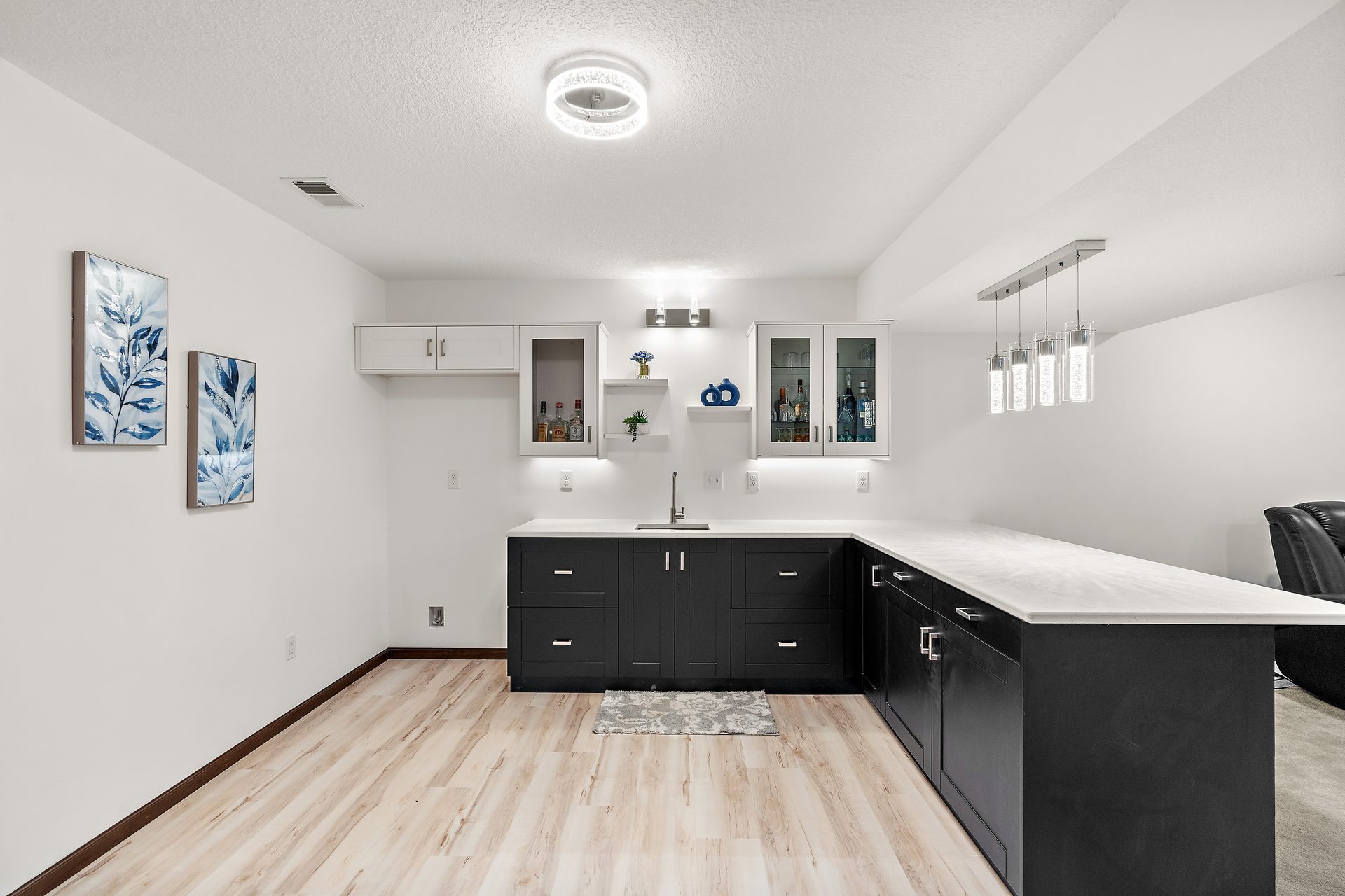 A basement wet bar with dark cabinetry, a quartz counter and pendant lighting over light plank flooring