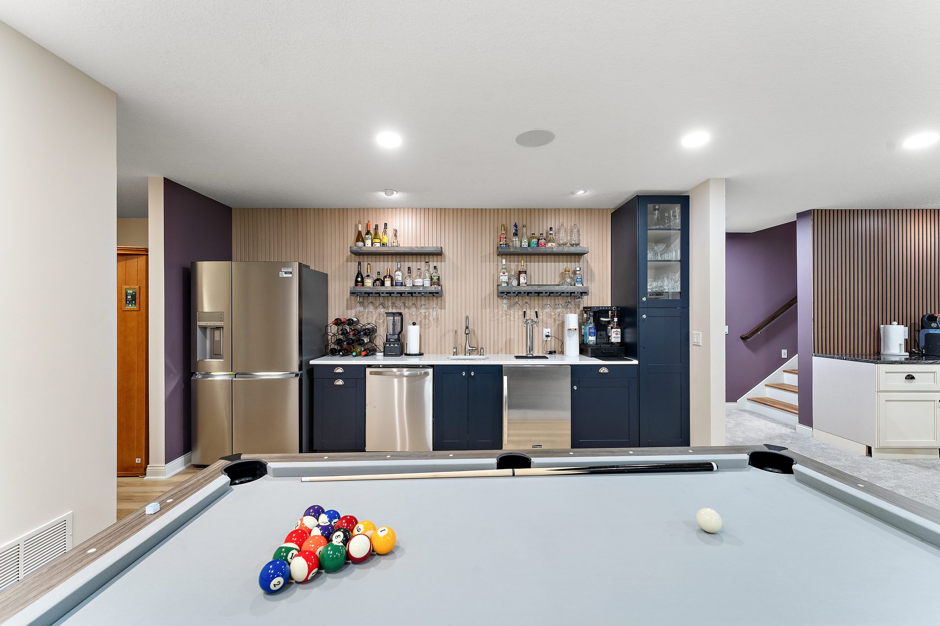A finished basement games room with a pool table, a navy wet bar and open shelving beneath a slat feature wall