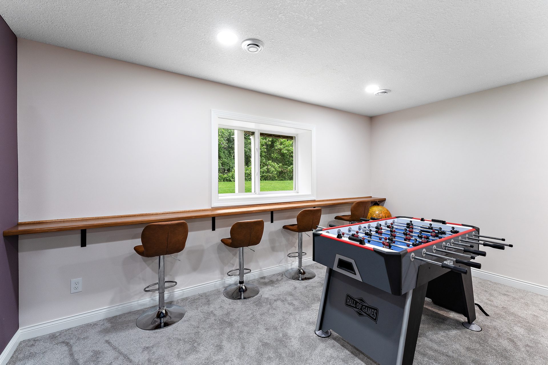 A finished basement games area with a rail bar and stools along the wall, beside a foosball table on carpet