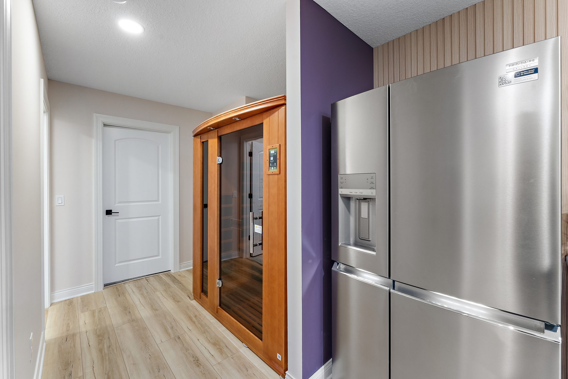 A cedar and glass sauna cabin built into a finished basement, beside a stainless refrigerator