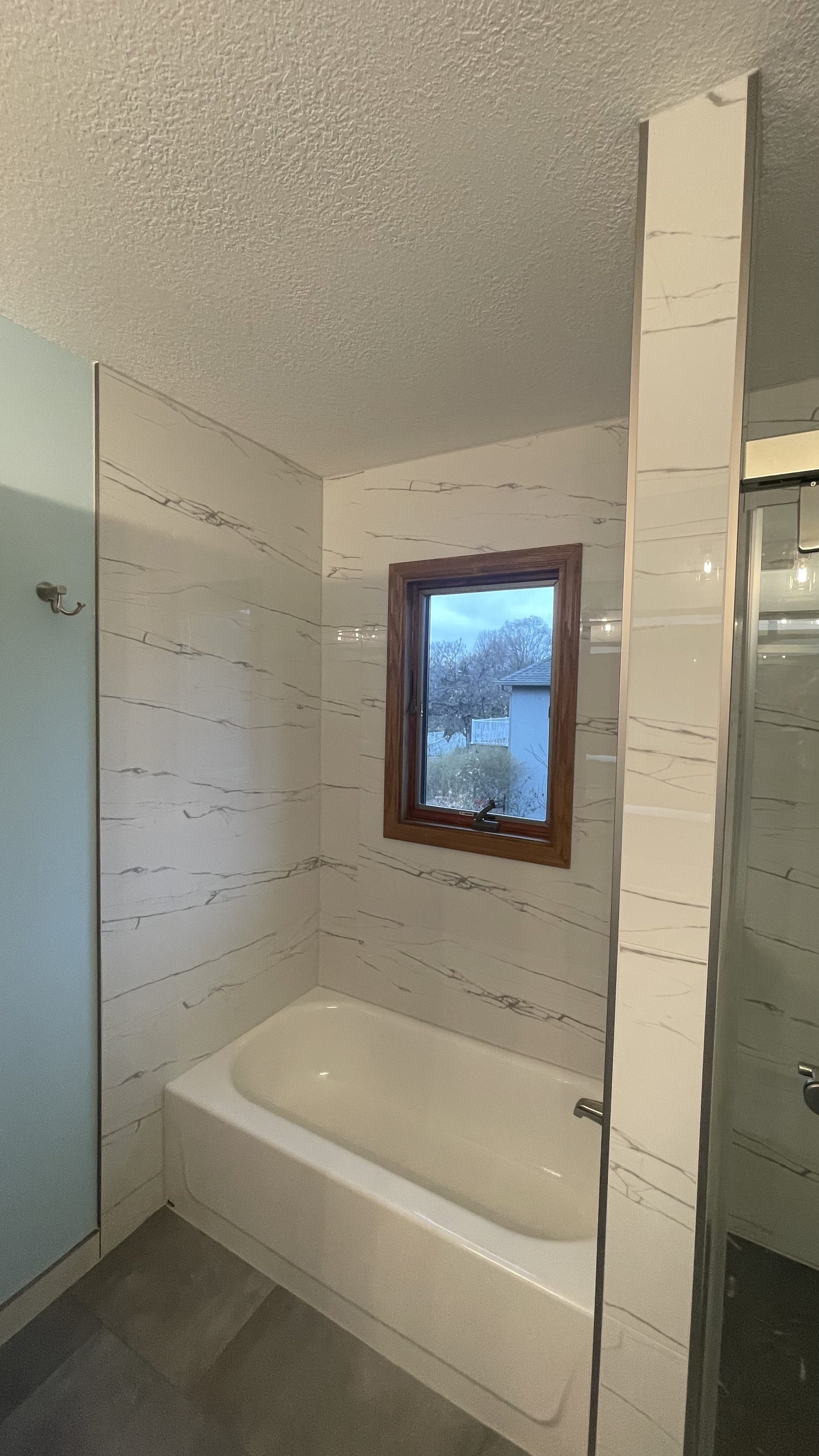 Tub and shower with a marble-look tile surround and a glass panel beside a trimmed window