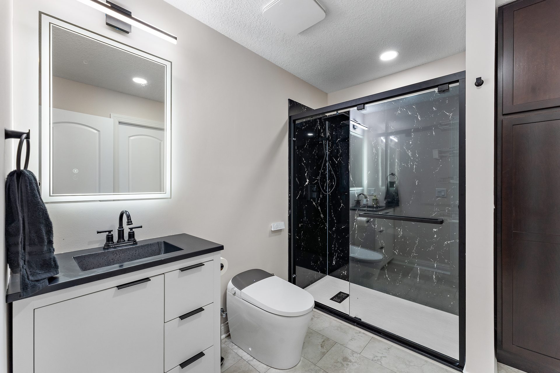 Bathroom with a white vanity, a black quartz top, a backlit mirror and a glass shower enclosure in black marble-look tile