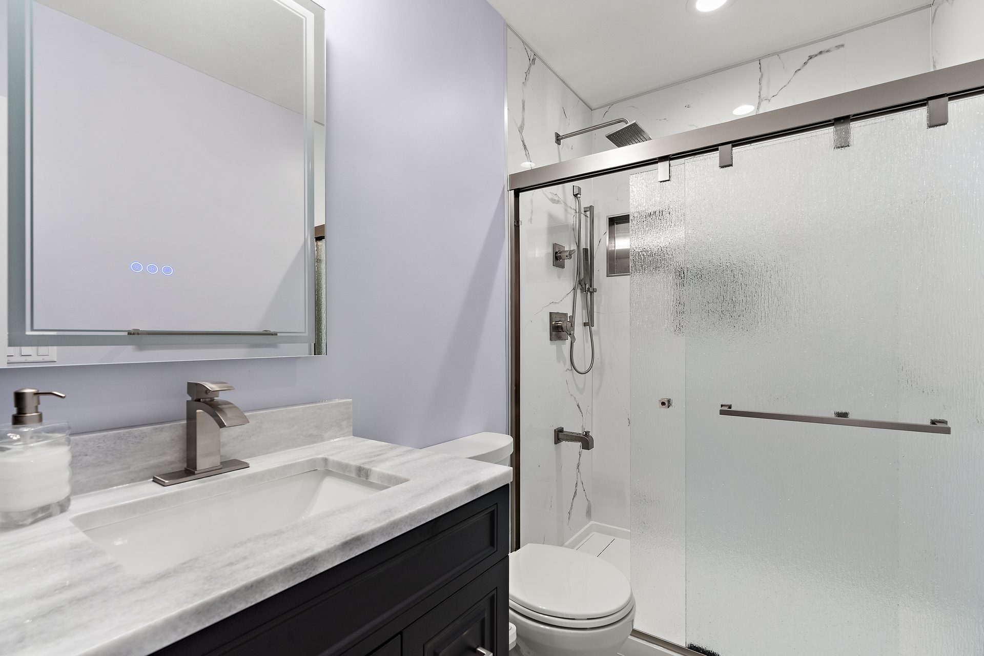 A bathroom with a dark vanity, a marble-look quartz top, a backlit mirror and a glass shower enclosure with a rain head