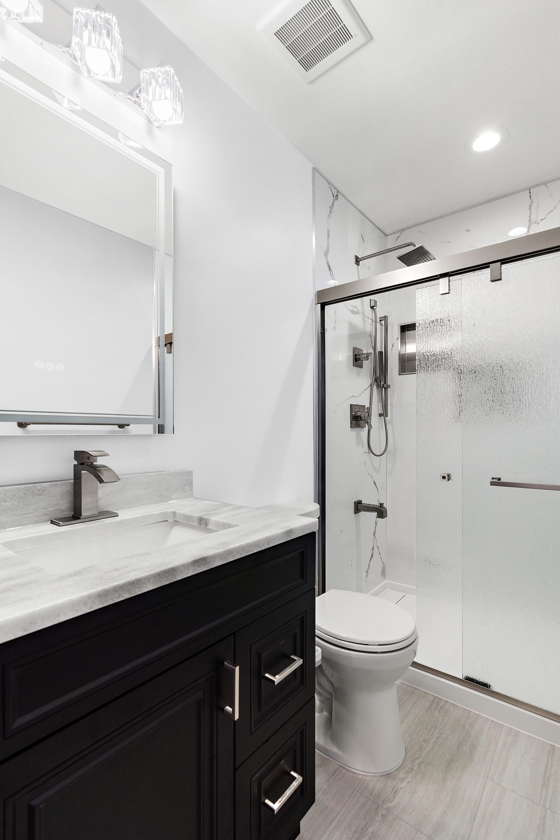 A bathroom with a dark vanity, a backlit mirror and a glass shower enclosure with a hand shower and rain head