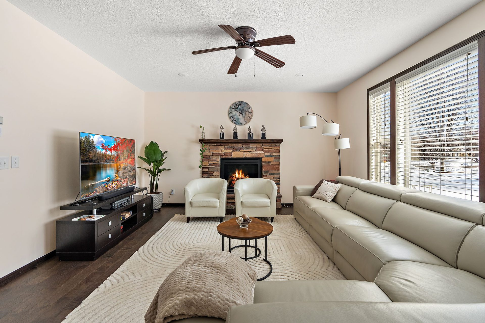 Living area with a stone fireplace surround, a ceiling fan and a wall of blinds