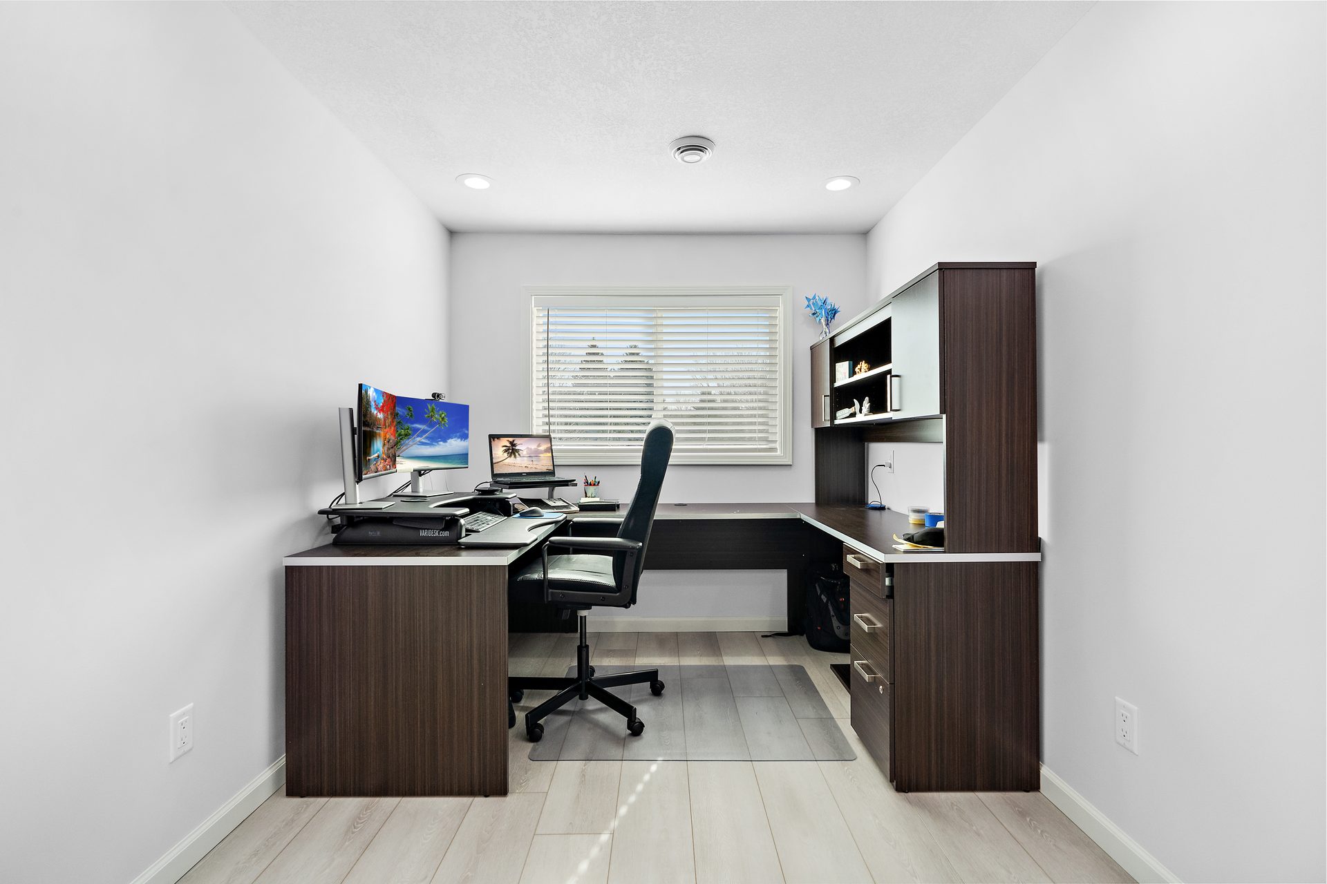 A built-in home office with a dark laminate desk run, upper storage and a blind-fitted window