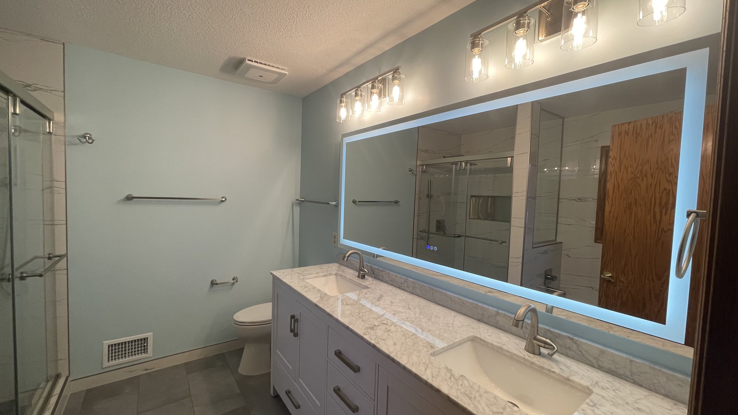 A remodeled bathroom with a double vanity, backlit mirror and tiled shower.